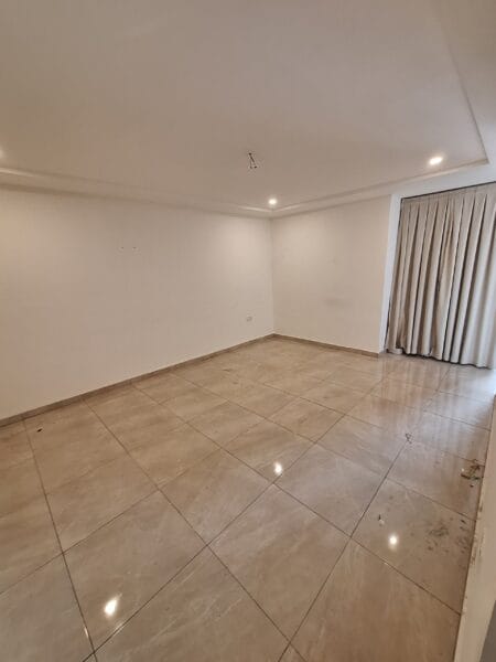 Empty living room with polished tiled floors, recessed lighting, and a large window covered by beige curtains.