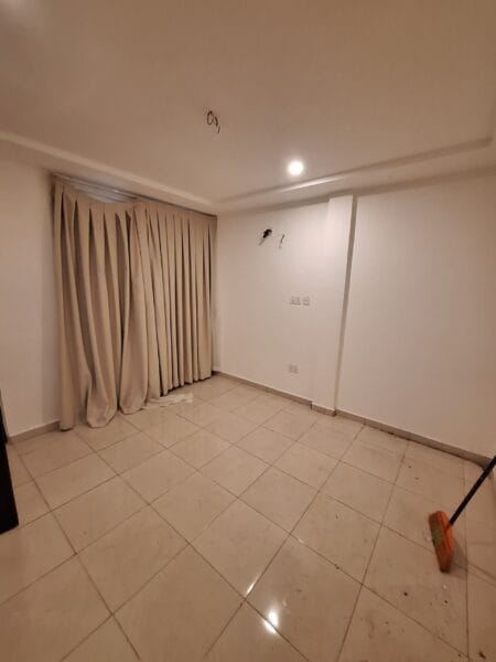 Empty living room with polished tiled floors, recessed lighting, and a large window covered by beige curtains.