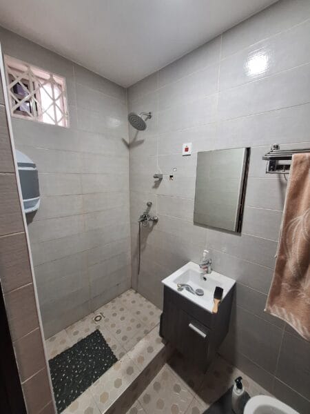 Modern bathroom with light gray tiles, walk-in shower, mirror, sink, and a small window for ventilation.