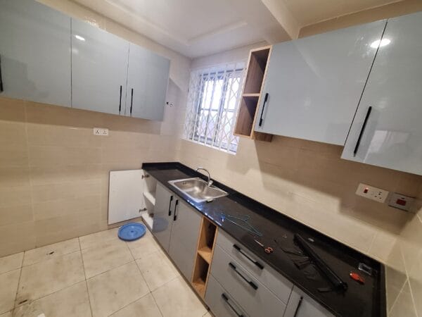 Compact kitchen with upper and lower cabinets, tiled backsplash, black countertop, and a stainless steel sink under a window.